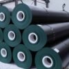 Black geomembrane rolls stacked horizontally in a storage area, with white cores and securing straps