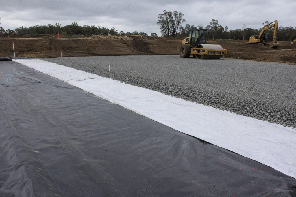 White geotextile and black geomembrane being rolled out over a prepared base on a road project