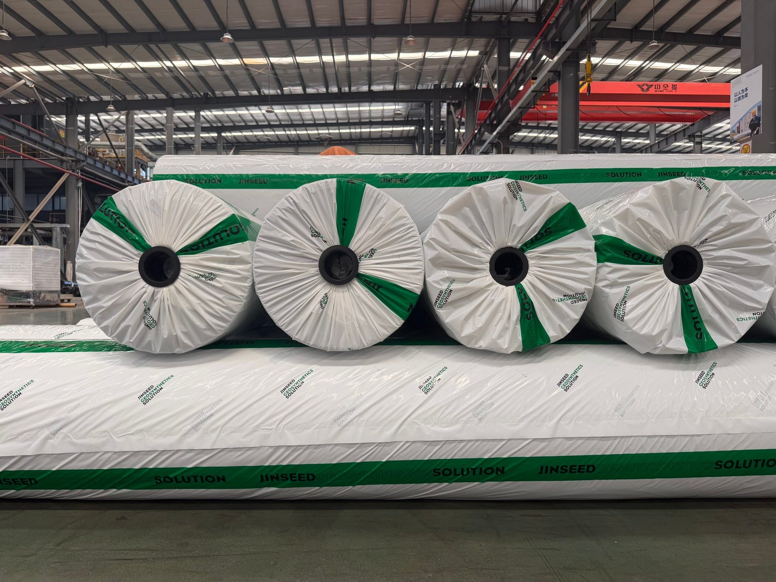 Geotextile rolls stacked in factory storage with exposed core ends and white-and-green protective wrapping