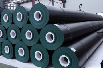 Black geomembrane rolls stacked horizontally in a storage area, with white cores and securing straps